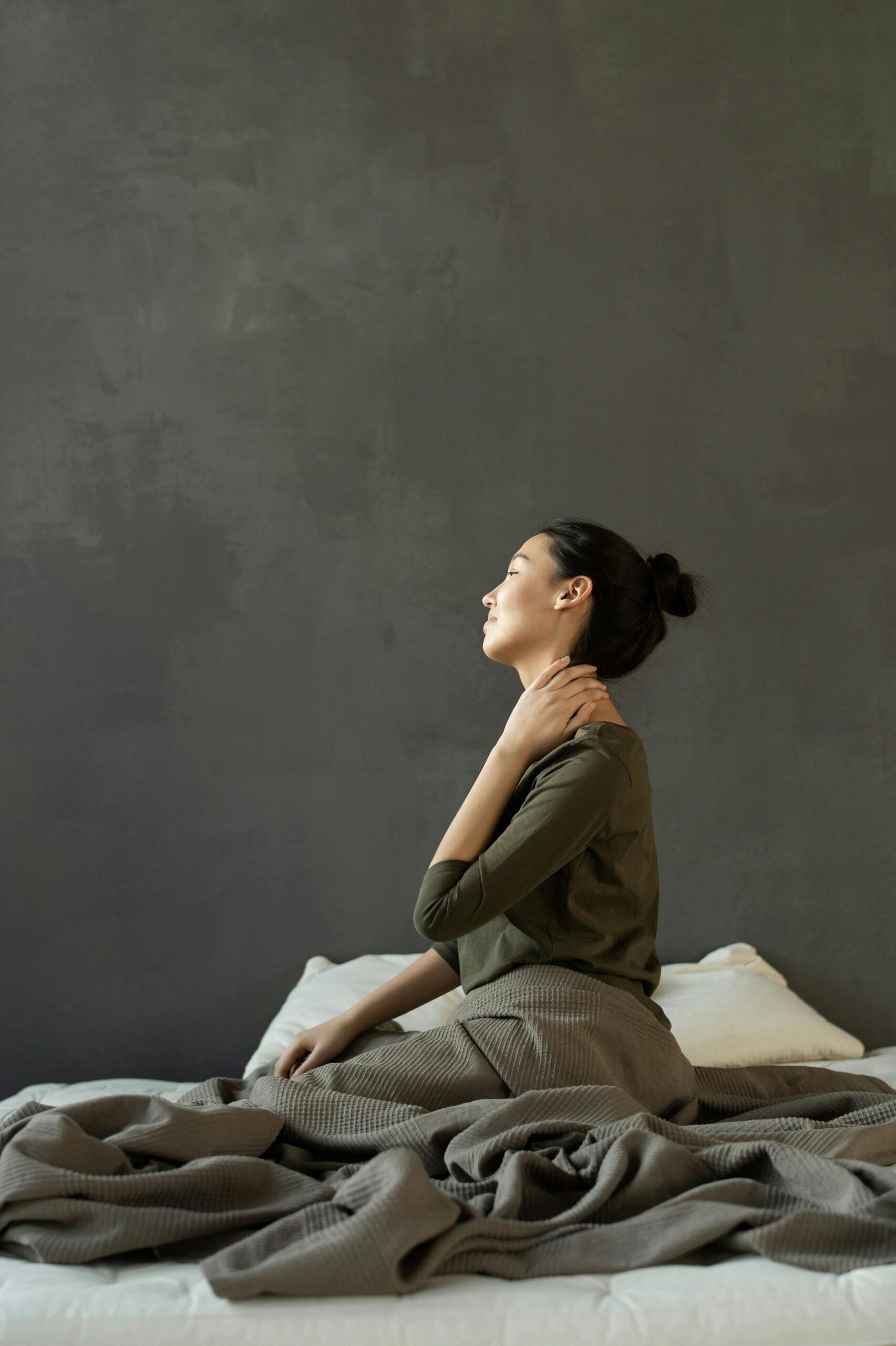 Side view of a woman sitting on a bed indoors, massaging her neck for stress relief.