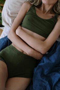 A woman in green clothing lies on a bed clutching her abdomen, suggesting pain or discomfort.