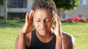 A woman holding her head in pain, indicating a severe headache outdoors in a sunny setting.