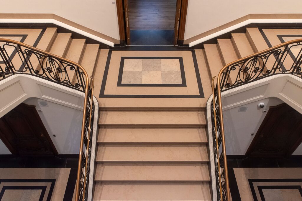 A top-down view of an ornate symmetrical staircase featuring intricate railings and patterned floors inside a classical building.